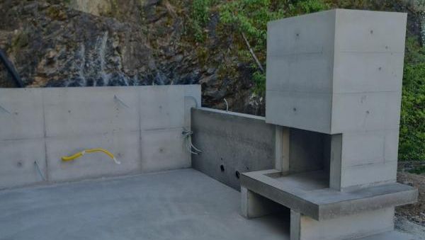 Utendørs betongstruktur med peis, plassert foran en steinvegg og omgitt av natur.